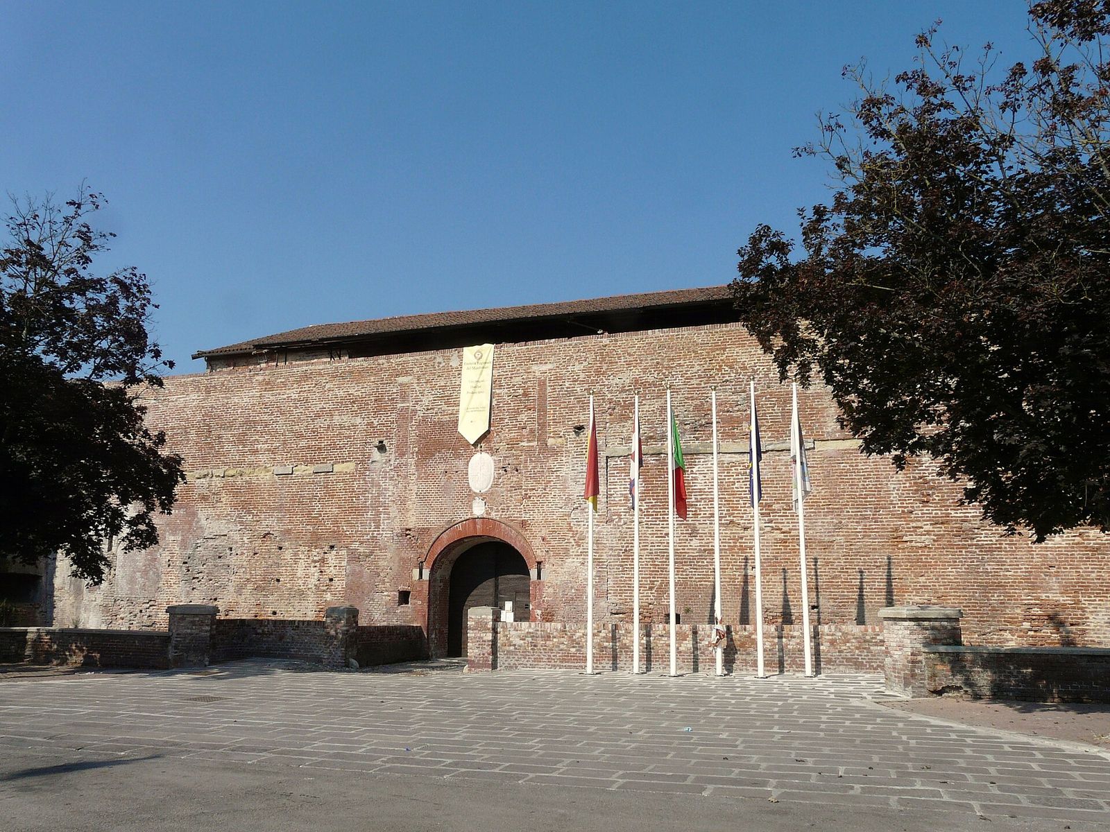 La facciata in mattoni del castello dei Paleologi a Casale Monferrato, con il portale d'ingresso e una fila di bandiere sotto il cielo azzurro