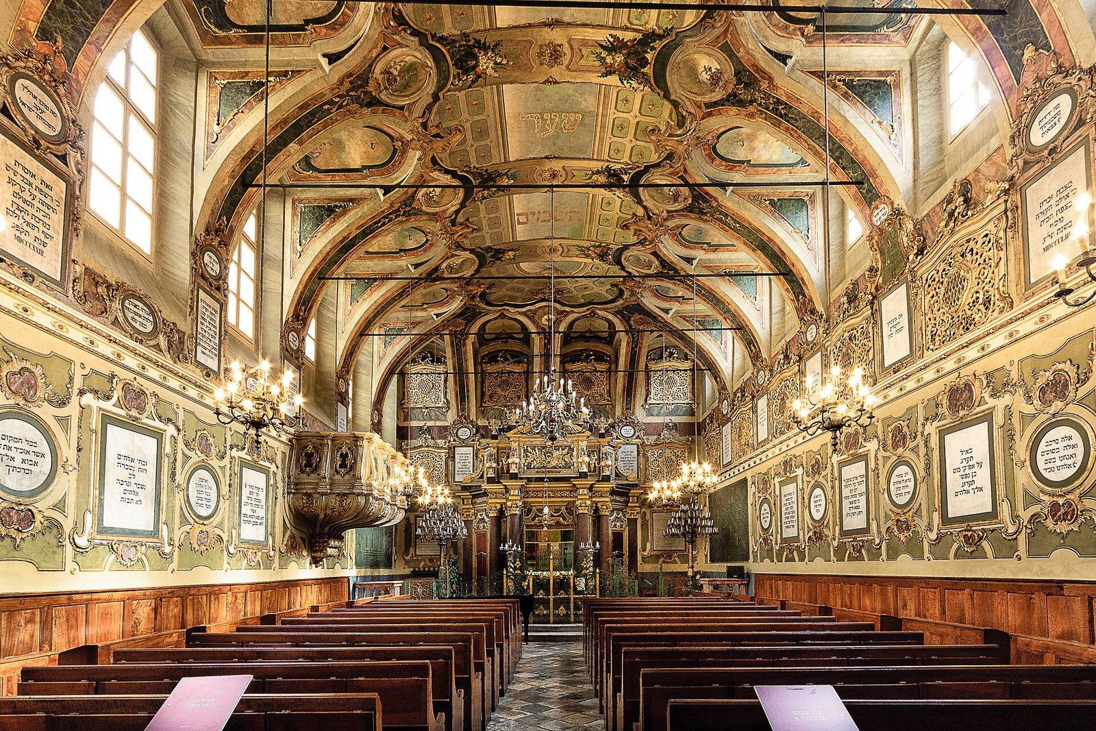 Interno barocco della sinagoga di Casale Monferrato con la volta dipinta, i lampadari accesi, le iscrizioni in ebraico e le panche in legno