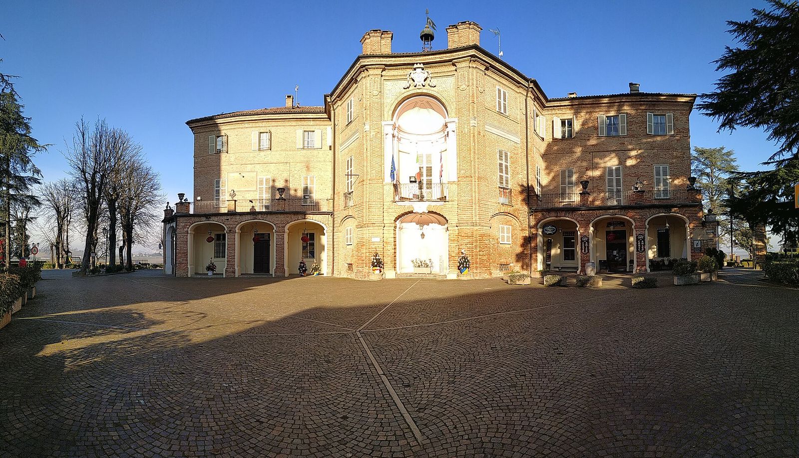 Facciata barocca in mattoni del castello di Castell'Alfero con i portici e la piazza lastricata, sotto un cielo azzurro