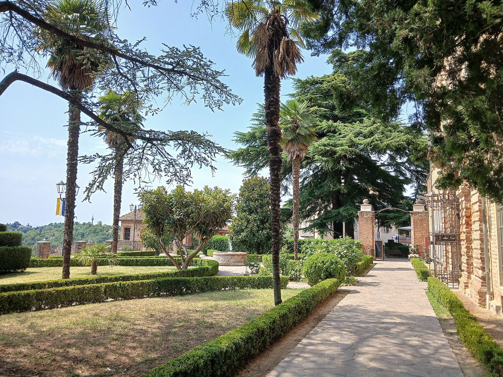 Giardini del castello di Castell'Alfero con palme, cedri, siepi di bosso e un viale lastricato accanto alla facciata in mattoni
