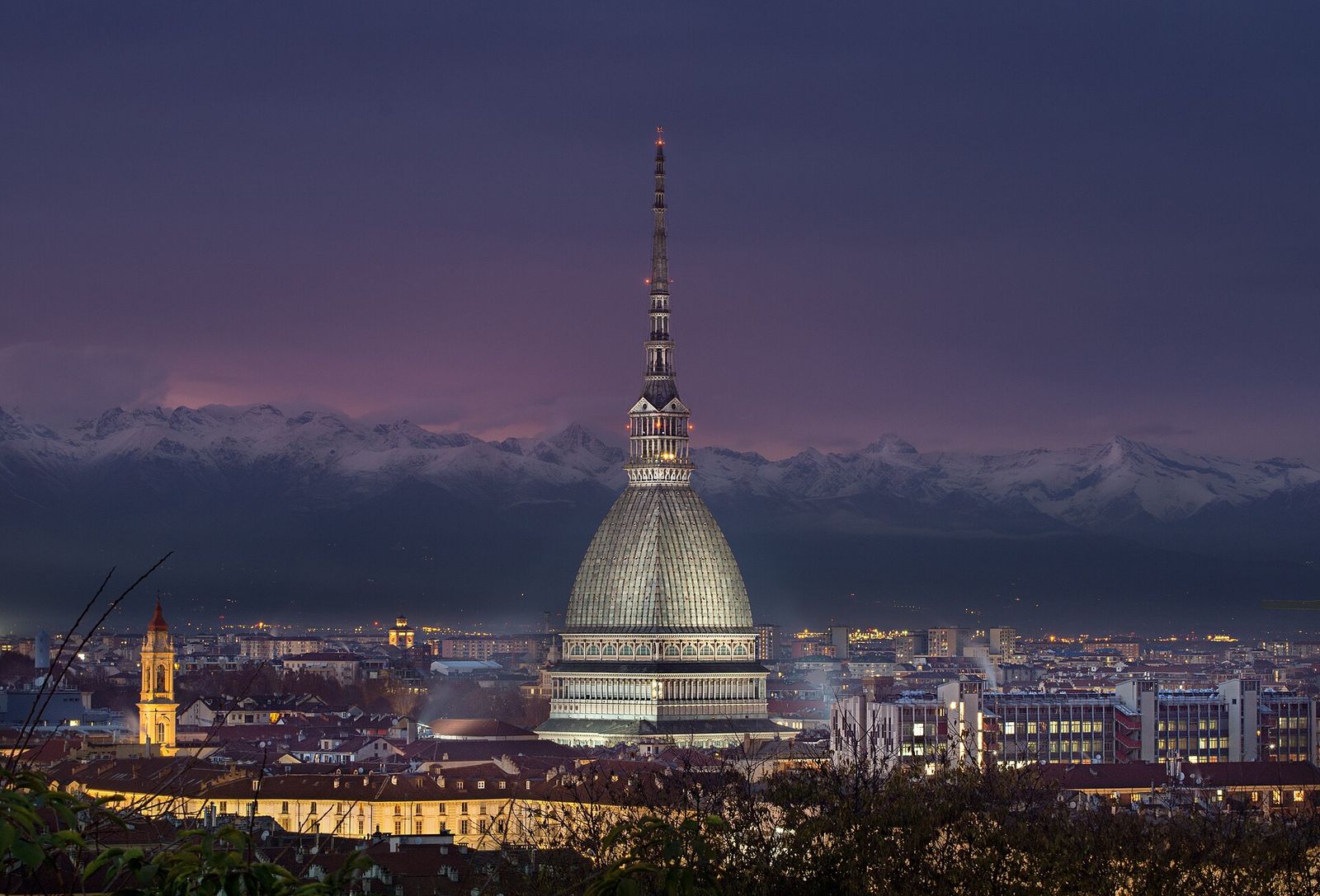 La Mole Antonelliana illuminata al crepuscolo sopra i tetti di Torino, con le Alpi innevate sullo sfondo